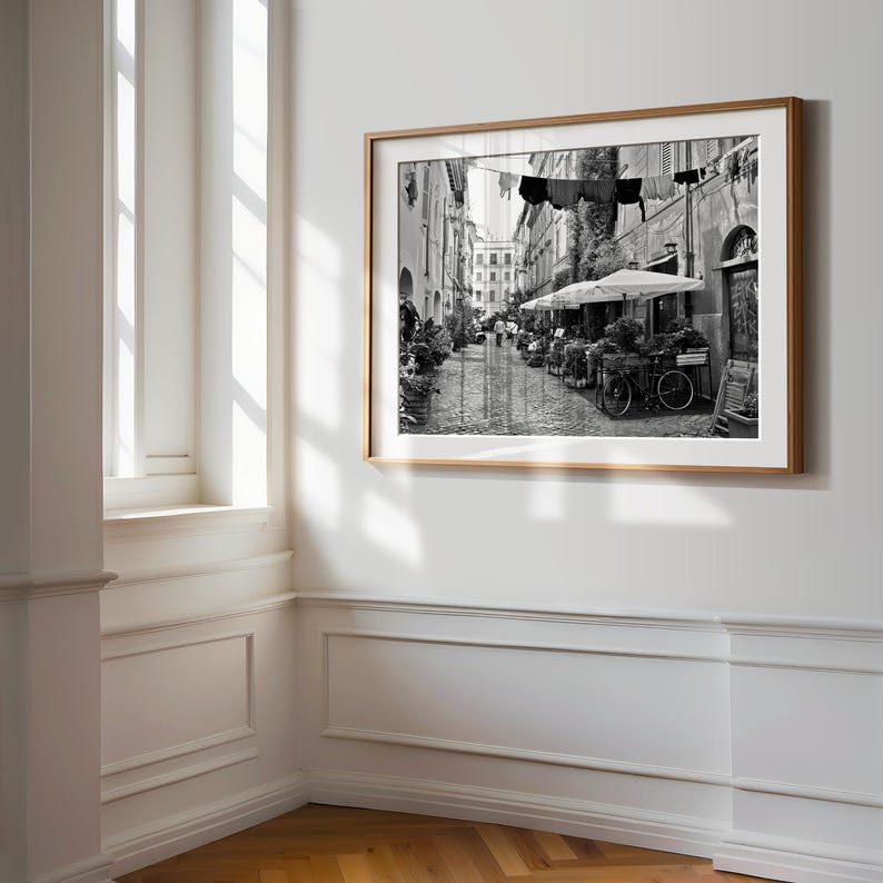 May include: A black and white framed wall art print depicting a narrow cobblestone street scene with laundry hanging on a line between buildings, outdoor cafe umbrellas, potted plants, and a bicycle parked along the side of the stone structures.