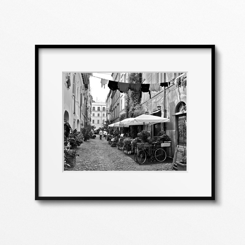 May include: A black and white framed photograph of a narrow cobblestone street lined with buildings. Laundry hangs on a line stretched across the alleyway above outdoor dining tables with white umbrellas and a bicycle parked near the wall.
