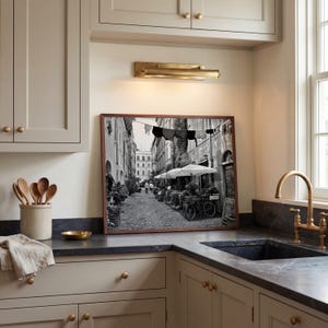 May include: A framed black and white photograph depicting a narrow European cobblestone street with laundry hanging on a line, outdoor umbrellas, and a bicycle, displayed on a kitchen counter beneath a brass wall light fixture.