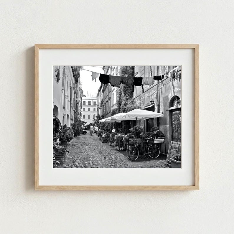May include: A black and white framed photograph depicting a narrow cobblestone street in Italy. The scene features laundry hanging on a line between buildings, outdoor cafe tables with white umbrellas, potted plants, and a bicycle parked nearby.