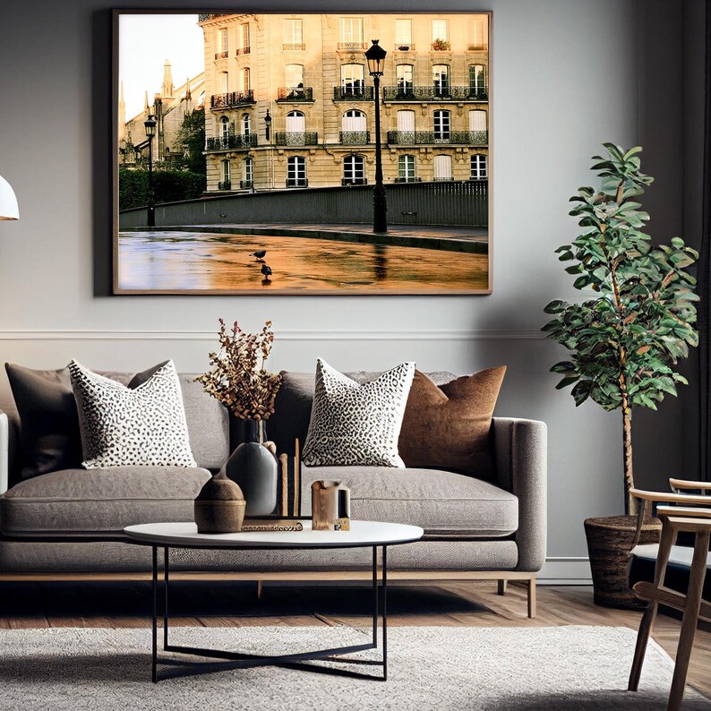 May include: A framed wall art print depicting a Parisian street scene with a stone building, wrought iron balconies, and a street lamp. The scene features a wet pavement reflecting warm light, with two small birds standing on the ground surface.