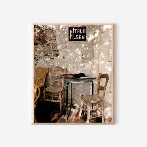 May include: A framed photograph of an outdoor cafe scene. The image features three wooden chairs, a small metal table, and a sign that reads "ITALA PILSEN". The background is a weathered stone wall.