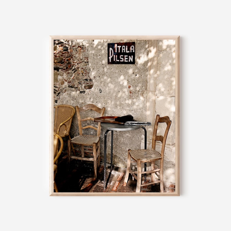 May include: A framed photograph of an outdoor cafe scene. The image features three wooden chairs, a small metal table, and a sign that reads "ITALA PILSEN". The background is a weathered stone wall.