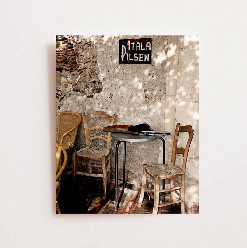 May include: A rustic cafe setting with wooden chairs and a small metal table. A sign reading "ITALA PILSEN" is mounted on a weathered stone wall. The scene is illuminated by sunlight, casting shadows.