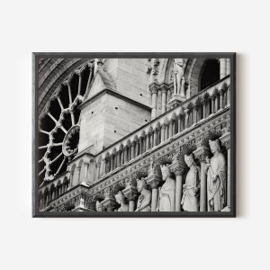 May include: A black and white framed print depicting the stone facade of Notre-Dame Cathedral. The image features a large circular rose window, a series of ornate stone columns, and a row of carved statues of figures standing in arched niches.