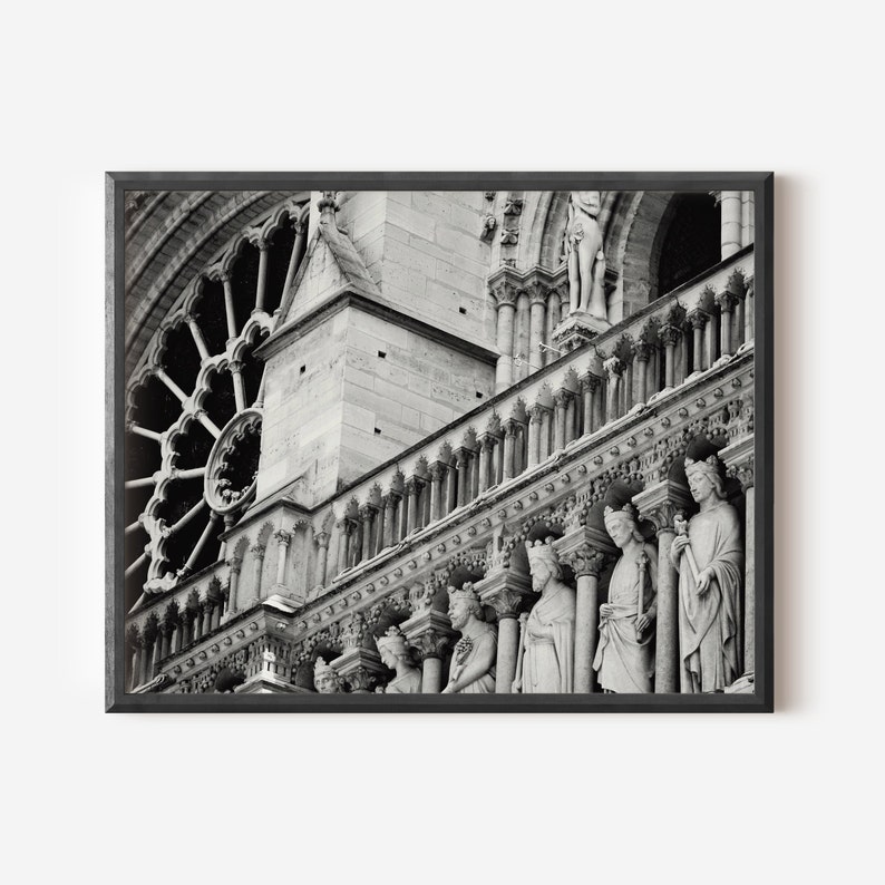May include: A black and white framed print depicting the stone facade of Notre-Dame Cathedral. The image features a large circular rose window, a series of ornate stone columns, and a row of carved statues of figures standing in arched niches.
