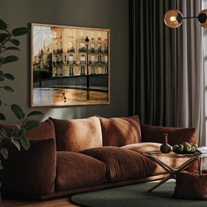 May include: A rectangular framed wall art print featuring a Parisian street scene with a beige stone building, wrought iron balconies, and a street lamp. The ground reflects the warm golden light of the setting sun, with two small birds visible.