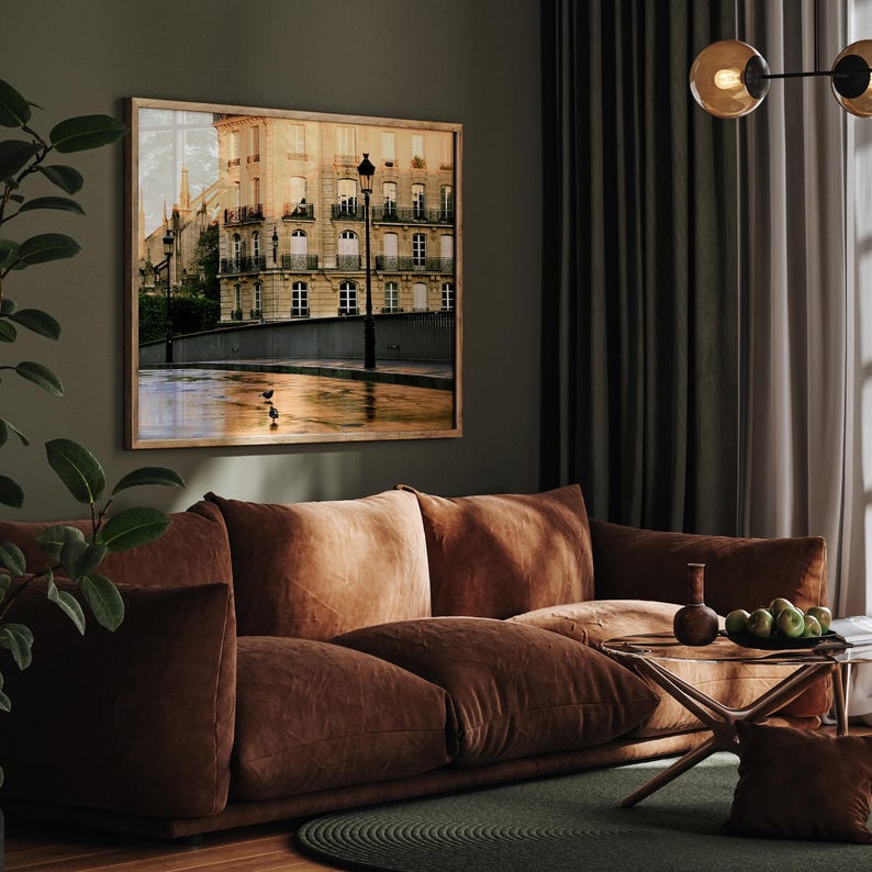 May include: A rectangular framed wall art print featuring a Parisian street scene with a beige stone building, wrought iron balconies, and a street lamp. The ground reflects the warm golden light of the setting sun, with two small birds visible.