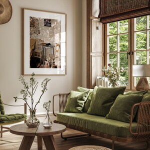 May include: A stylish living room interior with a framed picture, a green velvet sofa, and a wooden coffee table. The room is illuminated by natural light from a large window with a woven blind. Decorative vases with white flowers add a touch of elegance.