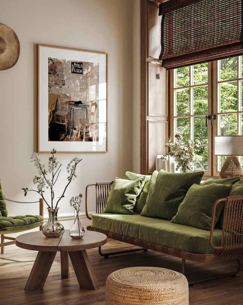 May include: A stylish living room interior with a framed picture, a green velvet sofa, and a wooden coffee table. The room is illuminated by natural light from a large window with a woven blind. Decorative vases with white flowers add a touch of elegance.