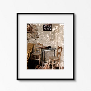 May include: A quaint outdoor cafe setting featuring wooden chairs and a small table. A sign reading "ITALA PILSEN" is mounted on a weathered wall. The scene is illuminated by sunlight, casting shadows.