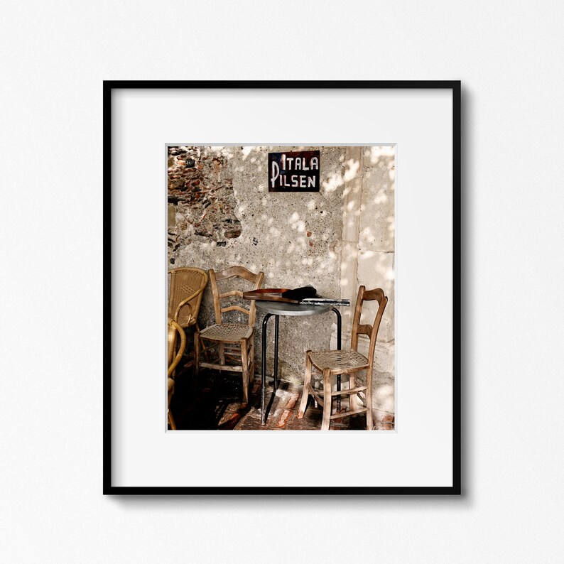 May include: A quaint outdoor cafe setting featuring wooden chairs and a small table. A sign reading "ITALA PILSEN" is mounted on a weathered wall. The scene is illuminated by sunlight, casting shadows.