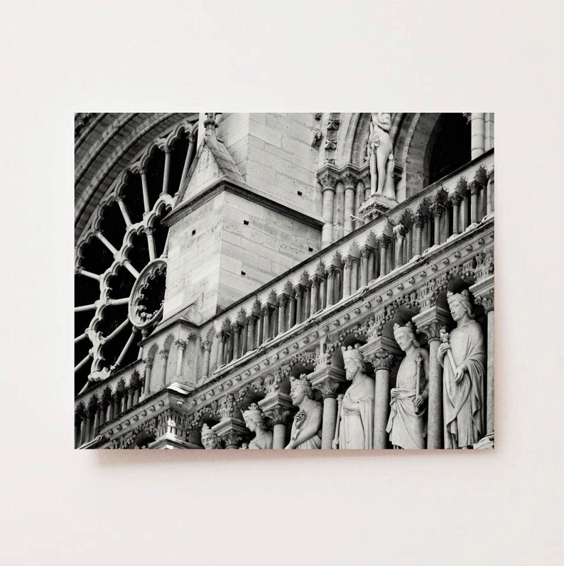 May include: A black and white architectural photograph of the Notre-Dame Cathedral facade, featuring a large circular rose window, stone columns, and a gallery of carved stone statues of figures wearing crowns, set against a plain white background.