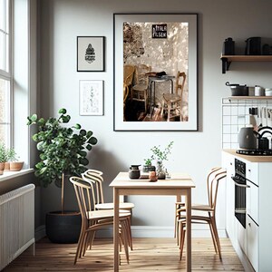 May include: A bright kitchen featuring a wooden table and chairs. A large framed print of a cafe scene hangs on the wall, alongside two smaller framed prints. A large potted plant and decorative items are also present.