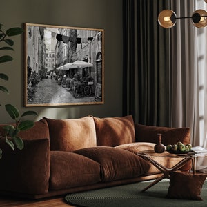 May include: A rectangular framed wall art piece featuring a black and white photograph of a narrow European street scene with cobblestones, outdoor cafe umbrellas, potted plants, and laundry hanging on a line between buildings.