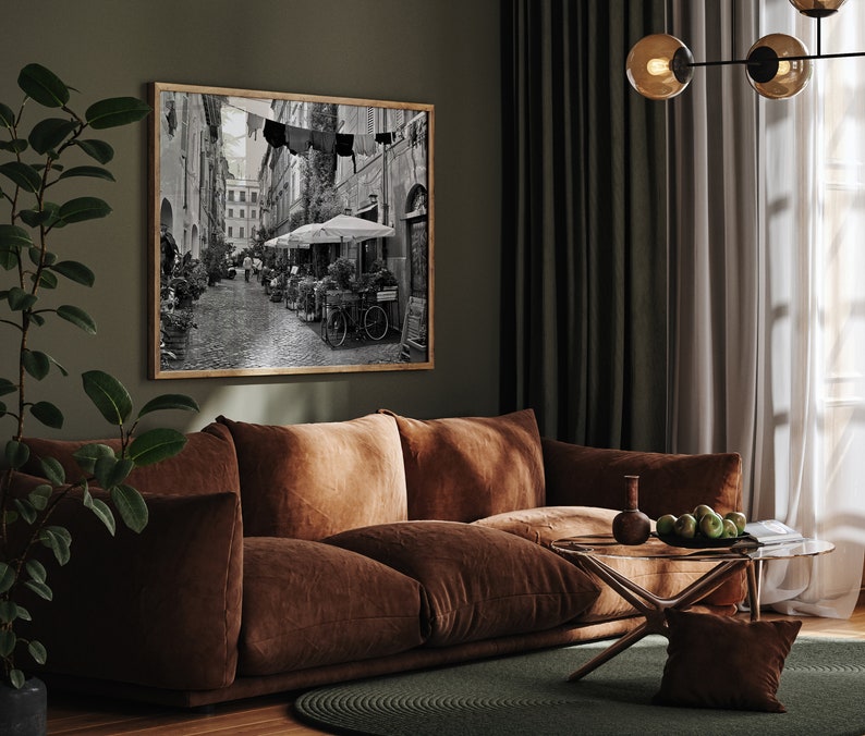 May include: A rectangular framed wall art piece featuring a black and white photograph of a narrow European street scene with cobblestones, outdoor cafe umbrellas, potted plants, and laundry hanging on a line between buildings.