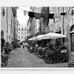 May include: A black and white photograph of a narrow cobblestone street in Rome, featuring laundry hanging on a line between buildings, outdoor dining umbrellas, potted plants, a bicycle, and graffiti on a wooden door in the background.