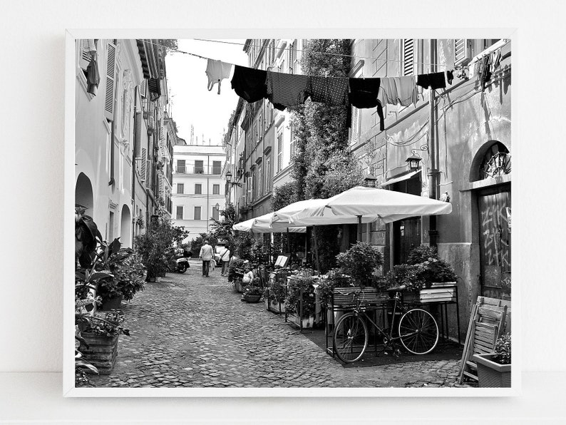 May include: A black and white photograph of a narrow cobblestone street in Rome, featuring laundry hanging on a line between buildings, outdoor dining umbrellas, potted plants, a bicycle, and graffiti on a wooden door in the background.