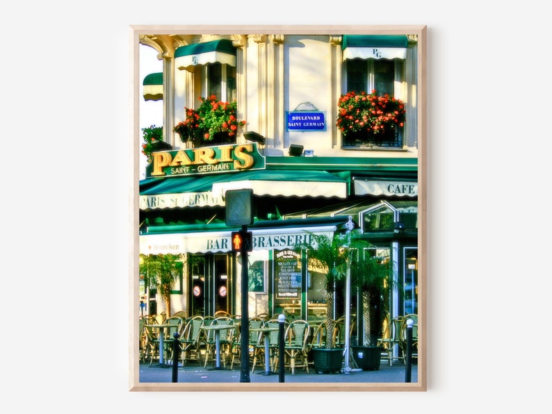 Vibrant colorful photography print of Paris Saint Germain cafe in the heart of Paris France.  Green awnings, a neon yellow sign and pops of red and pink from the flower boxes in the windows above  bistro tables and chairs lining sidewalk.