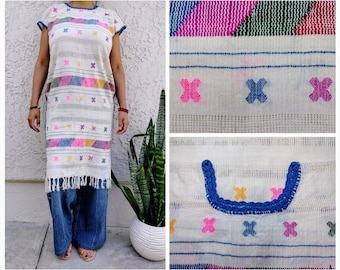 May include: A white tunic dress with colorful embroidered details. The dress features a fringe hem, short sleeves, and a round neckline with blue trim. The design includes horizontal stripes and cross-stitch patterns in pink, blue, yellow, and purple.