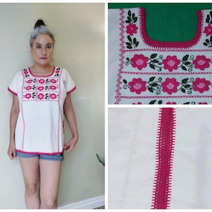 May include: White embroidered top with short sleeves and a square neckline. The top features pink floral embroidery along the neckline, chest, and hem. The edges are trimmed with pink stitching. A woman is wearing the top.