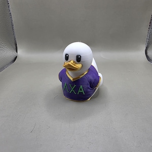 May include: A white duck figurine with a gold beak and black eyes. The duck is wearing a purple shirt with the letters ΛXA in green. The figurine is a novelty item.