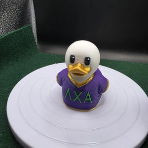 May include: A 3D-printed duck figurine with a white head, black eyes, and a gold beak. The duck wears a purple shirt with gold trim and green Greek letters. The figurine is on a white, circular surface.