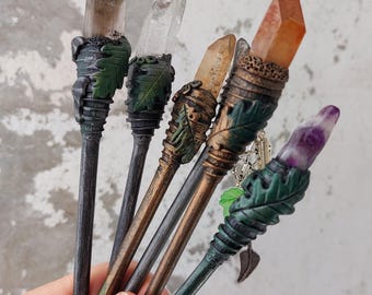 May include: A collection of handcrafted crystal wands. Each wand features a different colored crystal, including clear, brown, orange, and purple. The wands have dark wooden handles with decorative leaf and metal accents.