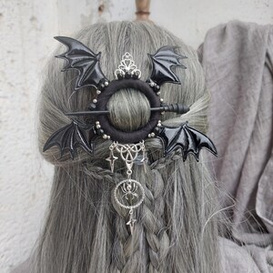 Può includere: Un accessorio per capelli nero con ali di pipistrello, un ciondolo a forma di ragno argentato e pendenti a stella. L'accessorio è posto su capelli grigi, con un anello nero e dettagli argentati. Design gotico o fantasy.