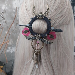 May include: A decorative hair accessory with a crescent moon, wings, and floral accents. The hair piece has a black circular base with a decorative pin, pink flowers, and dangling charms. The accessory is designed to be worn in the hair.