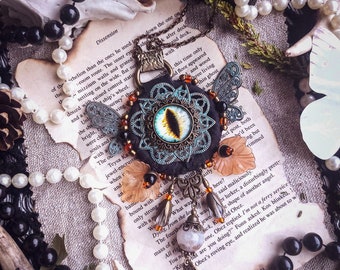 Puede incluir: Un collar con un gran colgante adornado con un ojo azul en un marco dorado, rodeado de intrincados trabajos en metal y cuentas de colores. El colgante está unido a un cordón negro con un cierre plateado. El collar está sobre un trozo de papel con texto sobre un viaje.