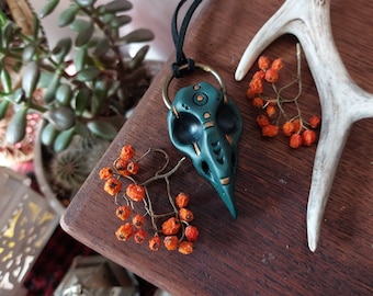 May include: A dark green ceramic bird skull pendant with gold accents and geometric patterns. The pendant is hanging from a black cord and is on a wooden surface with orange berries and a white antler.