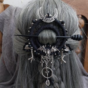 May include: A black hair accessory featuring a circular design with a crescent moon, bat, and star charms. The hair piece is adorned with black beads and a long, dark pin. The accessory is set against gray hair.