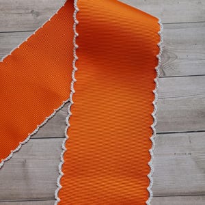 May include: Bright orange ribbon with a ribbed texture and a white, scalloped edge. The ribbon is folded over itself, showcasing its width and the contrasting white trim. This decorative ribbon is suitable for crafts and gift wrapping.
