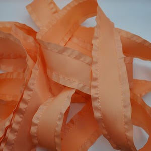 May include: Close-up of peach-colored ribbon with a stitched edge. The ribbon is folded and layered, creating a textured effect. The fabric has a slight sheen, and the stitching is visible along the edges.