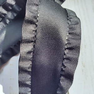 May include: Black satin ribbon with a ruffled edge. The ribbon is folded in half and the ruffled edge is visible on both sides.