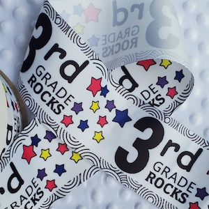 May include: White ribbon with black text that reads "3rd Grade Rocks" and black and white stars. The ribbon is folded in half.