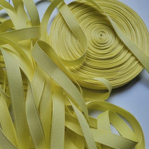 May include: A spool of pale yellow grosgrain ribbon, with loose lengths of the same ribbon scattered around. The ribbon has a ribbed texture and is likely used for crafting, sewing, or gift wrapping. The ribbon is a soft, pastel yellow.