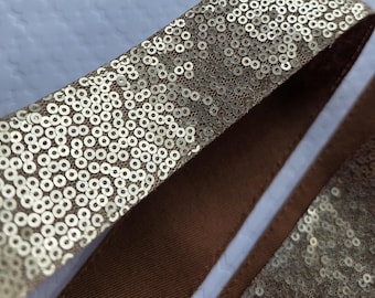 May include: A strip of gold sequin trim featuring a dense pattern of small, circular, metallic gold sequins attached to a brown fabric backing. The trim has a smooth, solid brown fabric edge on one side and a sequined surface on the other side.