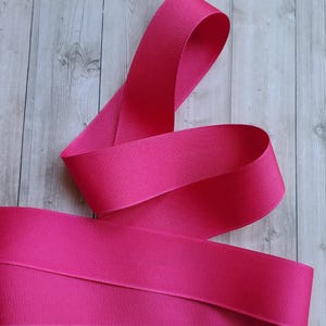 May include: Bright pink grosgrain ribbon, ideal for crafting and embellishments. The ribbon is wide, with a ribbed texture, and is shown in a flowing, curved arrangement against a wood grain background.
