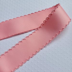 May include: A length of pink ribbon with a scalloped edge. The ribbon has a ribbed texture and a delicate, decorative trim. The ribbon is a soft, pastel pink color, suitable for crafts, gift wrapping, or embellishments.