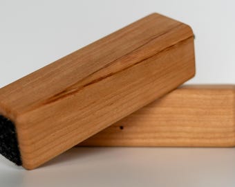May include: Two rectangular wooden erasers with a light brown finish. One eraser is resting on top of the other. The bottom of each eraser has a black felt pad for erasing. The wood grain is visible.