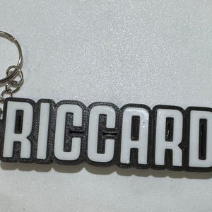 Op de afbeelding: Een sleutelhanger met de naam "RICCARDO" in witte blokletters met een zwarte omlijning. De sleutelhanger heeft een zilveren metalen ring en ketting bevestigd aan de naamplaat.