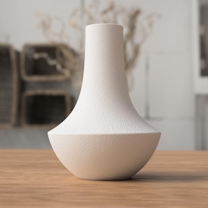 Può includere: Un vaso bianco, strutturato, dalla forma unica. Il vaso ha una base ampia e arrotondata che si assottiglia verso un collo stretto. La superficie ha una trama maculata. Il vaso è appoggiato su un tavolo di legno.