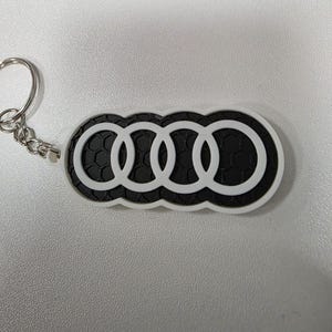 Könnte beinhalten: Ein schwarz-weißer Audi-Logo-Schlüsselanhänger. Der Schlüsselanhänger zeigt die ikonischen vier ineinandergreifenden Ringe in Weiß, vor einem schwarzen Hintergrund mit einem Wabenmuster. Der Schlüsselanhänger hat einen silbernen Metallring.