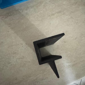 May include: A dark brown, L-shaped wooden bracket with a single, pointed support leg. The bracket is positioned on a light-coloured surface, with a blue object visible in the background. The bracket appears to be new.
