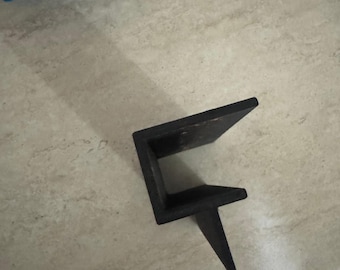 May include: A dark brown, L-shaped wooden bracket with a single, pointed support leg. The bracket is positioned on a light-colored surface, with a blue object visible in the background. The bracket appears to be new.