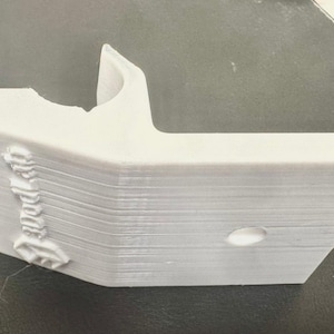 May include: A light gray, 3D-printed object with a unique shape. It features a curved section and a right-angled extension. The object has a hole and the word "Tool" is printed on the side. The item is made of layered material.