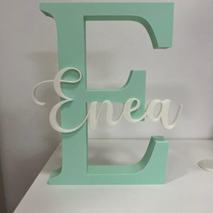 May include: A large, mint green, three-dimensional capital letter E with a smaller, white, cursive script name Enea layered across the center. The object is freestanding with a flat base and features a smooth, matte finish on all visible surfaces.