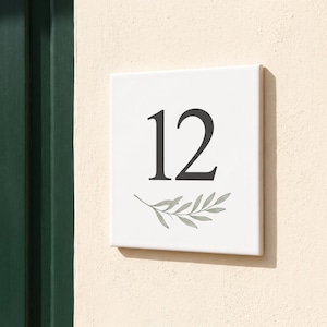 Könnte beinhalten: Ein quadratisches, weißes Hausnummernschild mit der Zahl 12 in einer dunkelgrauen Serifenschrift. Unter der Zahl befindet sich eine zarte Illustration eines grünen Olivenzweigs mit Blättern. Das Schild hat eine glatte Oberfläche.